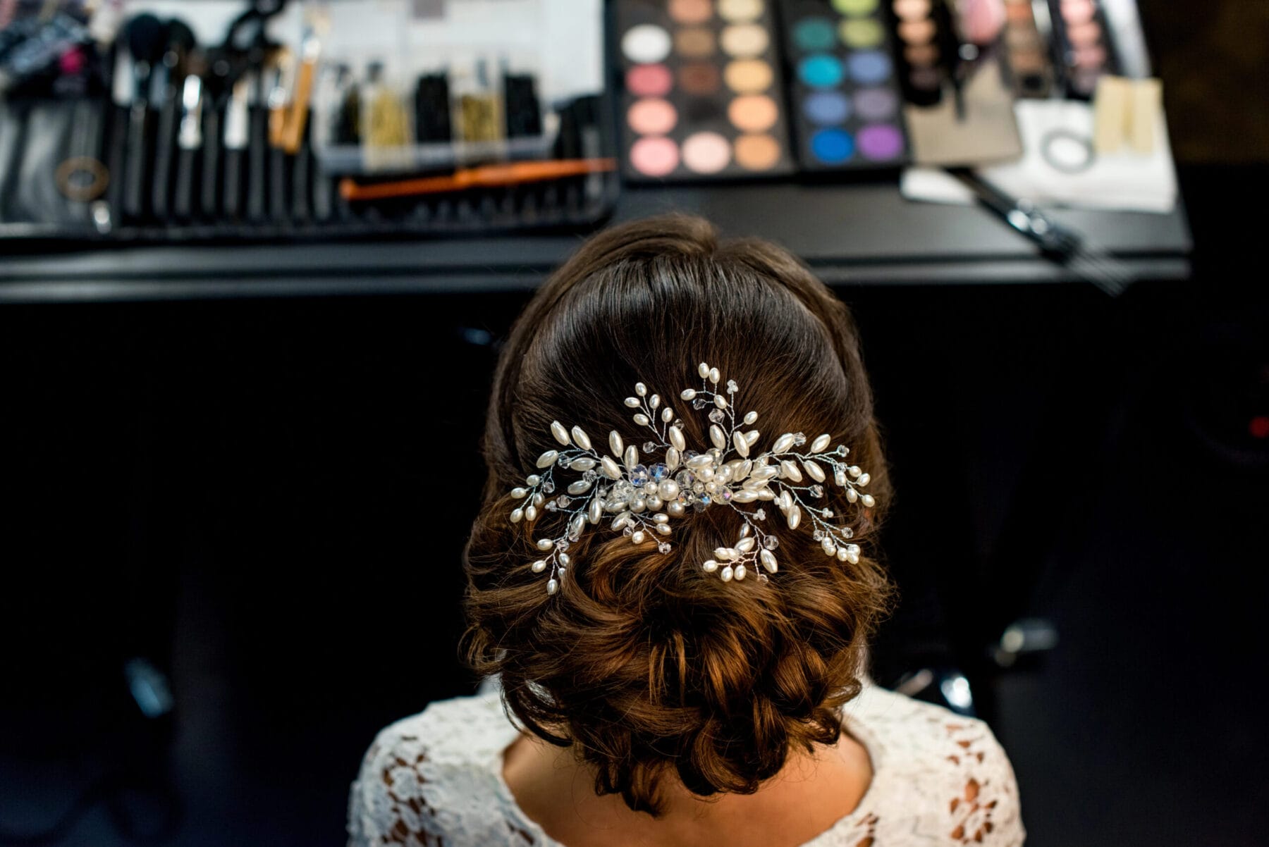 Experience relaxation and rejuvenation at YOLOve Beauty Spa in Dartmouth. A woman with intricately styled brown hair adorned with elegant pearls and silver leaves enjoys the serene ambiance. The makeup table, displaying a vivid eyeshadow palette and brushes, reflects the high-quality beauty services offered. Dressed in a white lace top, she epitomizes the elegance and transformative benefits of visiting this exquisite spa.